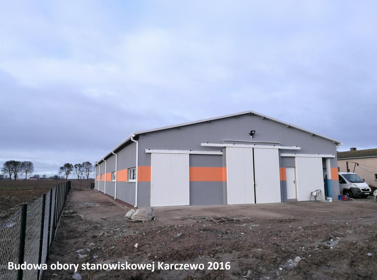 Szara hala z przesuwanymi białymi drzwiami i pomarańczowymi pasami, widoczny teren budowy, zaparkowany bus i częściowo widoczne drzewa w tle.