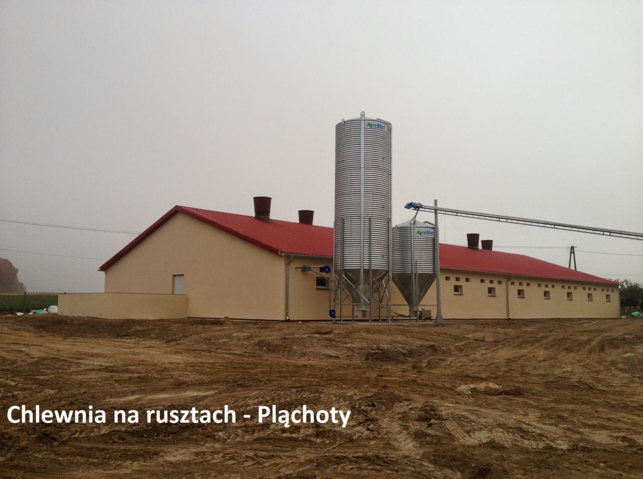 Nowoczesna chlewnia z czerwonym dachem i dwoma silosami zbożowymi AgroMat, widok z zewnątrz na tle pochmurnego nieba.