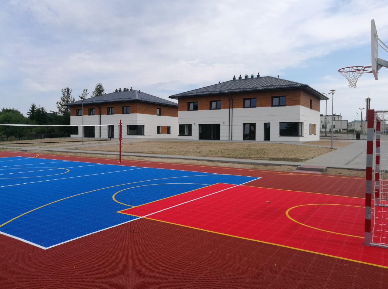 Dwa identyczne, nowo wybudowane budynki z drewnianą elewacją na tle boiska sportowego z siatką do siatkówki i koszem do koszykówki.