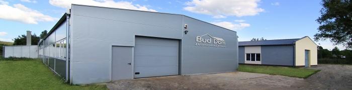 Szara hala przemysłowa z blachy trapezowej z bramą segmentową i drzwiami wejściowymi, widoczne logo firmy Bud Dom na elewacji, w tle mniejszy budynek.