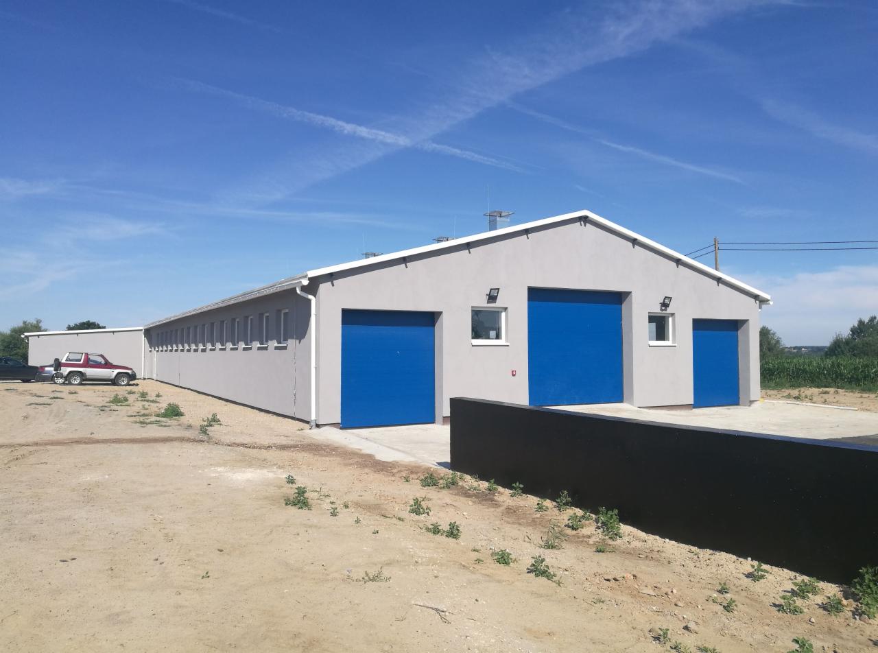 Szara hala z niebieskimi bramami segmentowymi, widoczne okna i oświetlenie zewnętrzne, na pierwszym planie czarny murek, w tle zaparkowany czerwony samochód terenowy.