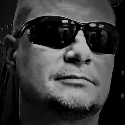 Czarno-biały portret mężczyzny w okularach przeciwsłonecznych, z krótkimi włosami i zarostem, patrzącego wprost, z łańcuszkiem na szyi.