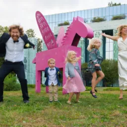 Radosna rodzina skacze na trawie przed różowym, drewnianym pegazem na tle nowoczesnego budynku z zielonymi akcentami.