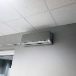 Klimatyzator ścienny, srebrny, zamontowany w biurze na szarej ścianie pod sufitem kasetonowym, widok z dołu.