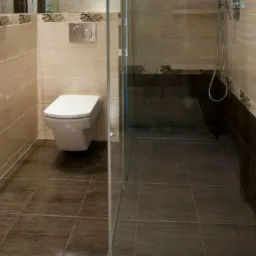 Nowoczesna łazienka z beżowymi i brązowymi płytkami, podwieszana toaleta z chromowanym przyciskiem spłukiwania, kabina prysznicowa ze szklaną ścianką.