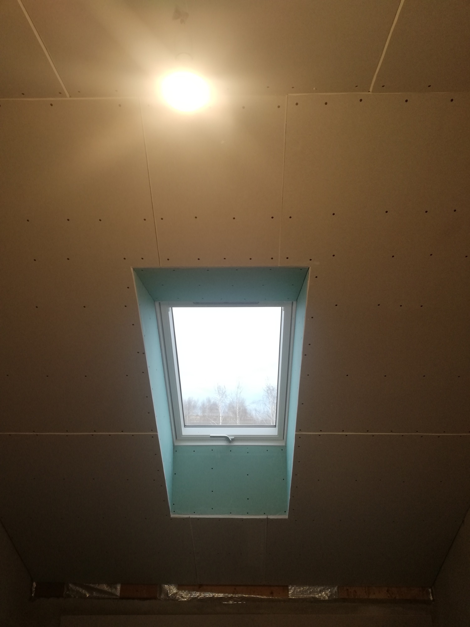 Montaż okna dachowego w suficie z płyty karton gips, widoczne łączenia płyt i wkręty, surowy stan wykończenia.