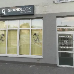 Fasada salonu z szyldem 'GRANDLOOK Salon fryzjerski', okna z grafikami kobiecych twarzy i godziny otwarcia na drzwiach wejściowych.