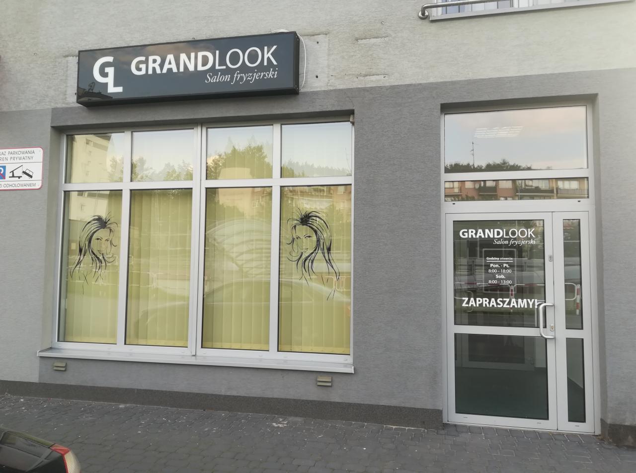 Fasada salonu z szyldem 'GRANDLOOK Salon fryzjerski', okna z grafikami kobiecych twarzy i godziny otwarcia na drzwiach wejściowych.