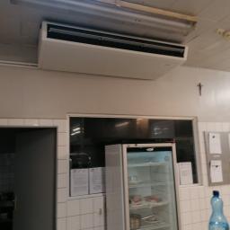 InstalCold - Sufitowy klimatyzator w pomieszczeniu z chłodziarką i kafelkami, widoczne oświetlenie sufitowe i krzyż na ścianie.