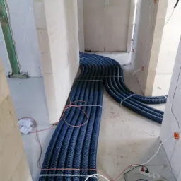 Rury wentylacyjne w korytarzu budynku w trakcie budowy, zabezpieczone taśmą, widoczne puszki elektryczne i przewody.