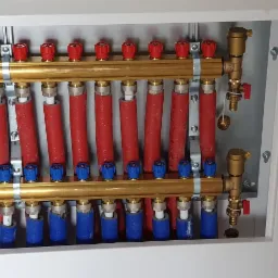 Rozdzielacz ogrzewania podłogowego w białej szafce, z czerwonymi i niebieskimi rurami oraz mosiężnymi elementami, widoczne zawory i odpowietrzniki.