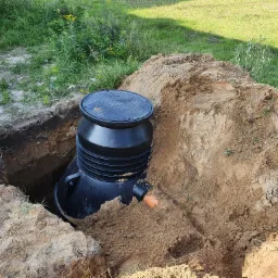 Czarny zbiornik z pokrywą, prawdopodobnie szambo lub oczyszczalnia, umieszczony w wykopie z ziemi.