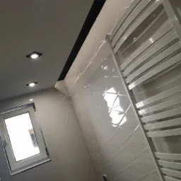 Biała łazienka z błyszczącymi, trójwymiarowymi płytkami ściennymi ułożonymi w romby, grzejnik drabinkowy i okno z białymi ramami.