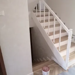 Klatka schodowa w Kalinowie po odświeżeniu: białe balustrady, drewniane stopnie, ściana pokryta dekoracyjną szpachlą, widoczne wiadro z farbą i zabezpieczona podłoga.