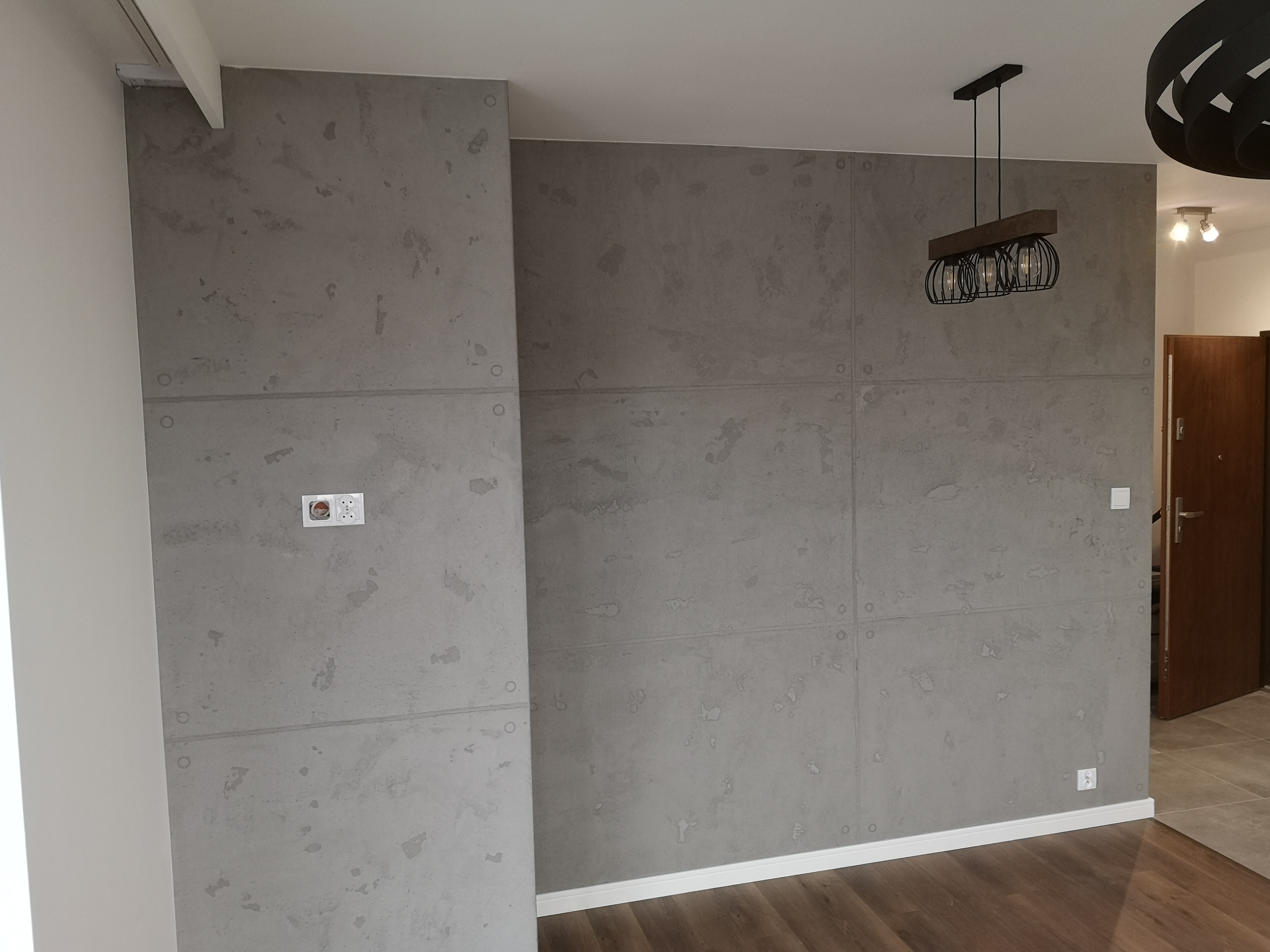 Ściana wykończona panelami imitującymi beton architektoniczny, z widocznymi gniazdkami elektrycznymi i ozdobnym oświetleniem sufitowym, uzupełnona drewnianą podłogą.