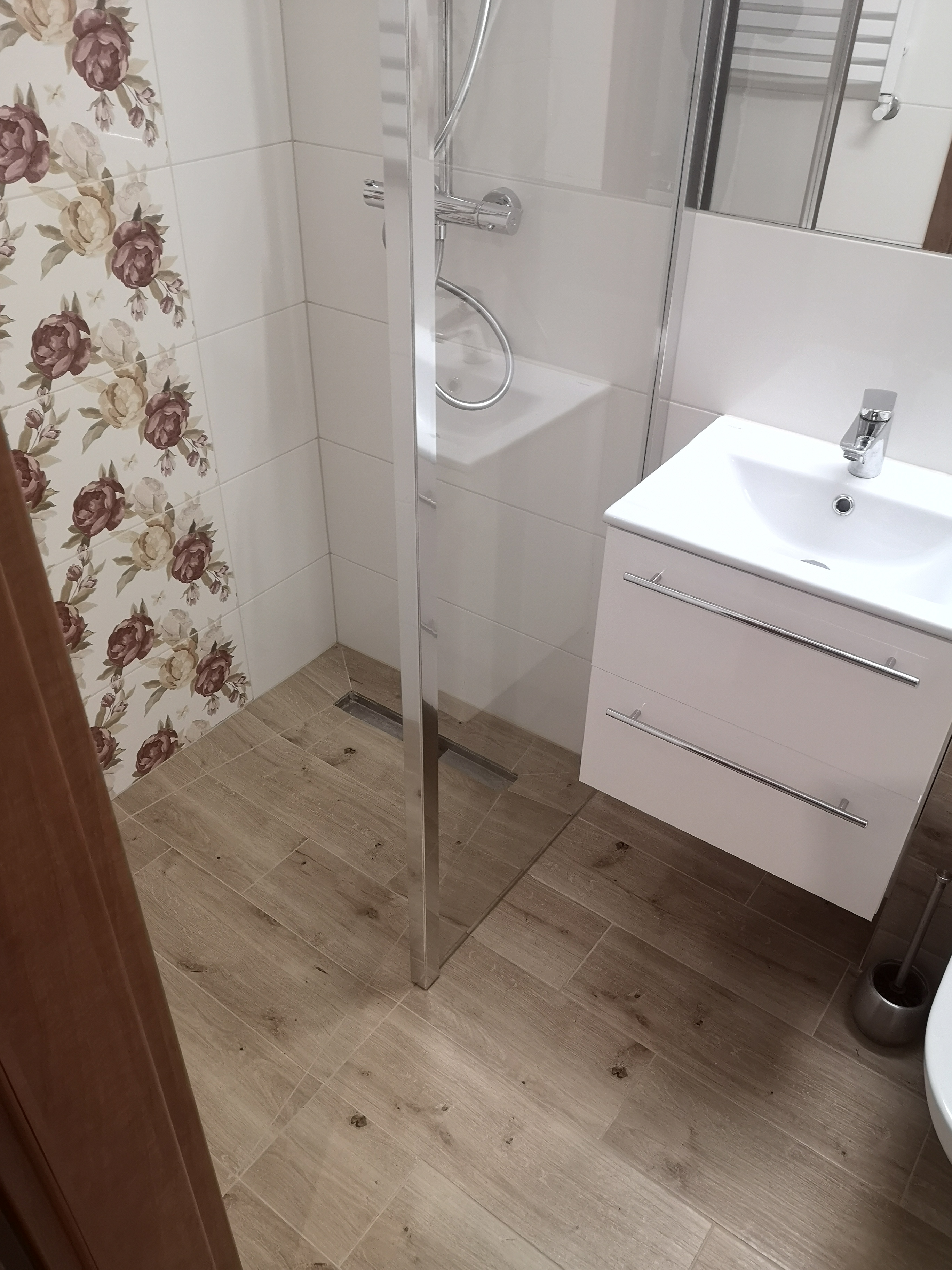 Ukończona łazienka z kabiną prysznicową ze szklaną ścianką, białą szafką z umywalką i baterią oraz płytkami podłogowymi imitującymi drewno, uzupełniona o dekoracyjne płytki ścienne z motywem...