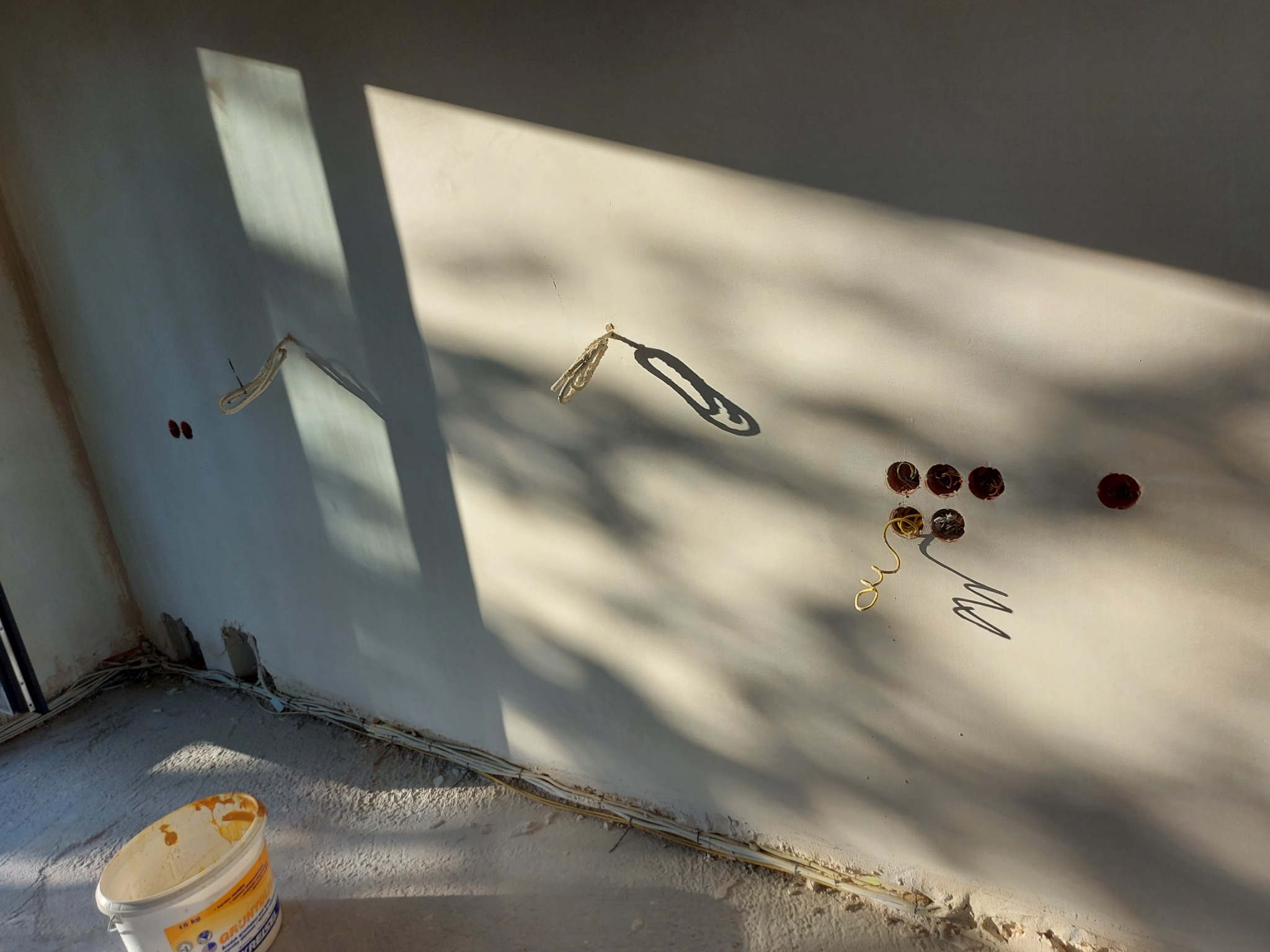 Ściana po nałożeniu tynku maszynowego, widoczne kable elektryczne wystające ze ściany, puszki instalacyjne, wiadro z gruntem i ostre światło słoneczne tworzące wyraźne cienie.