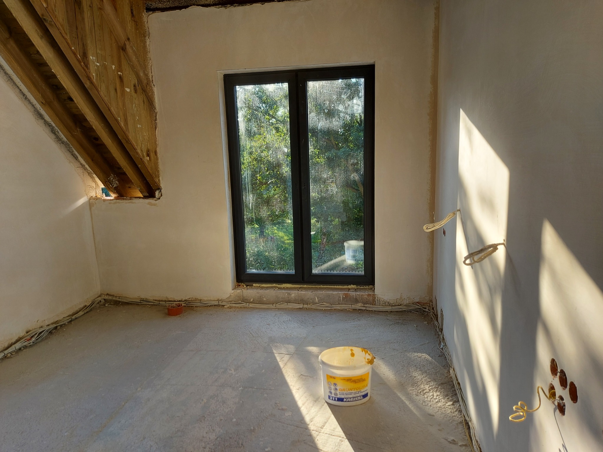 Wnętrze pokoju po nałożeniu tynków, widoczne przewody elektryczne wystające ze ścian, okno z widokiem na zieleń oraz wiadro z gruntem podłogowym.