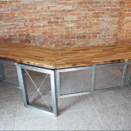 Narożny blat z drewna klejonego, z ciemnymi krawędziami, na metalowej konstrukcji z siatką, na tle ceglanej ściany.