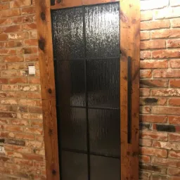 Drewniane drzwi przesuwne w stylu loft z czarną ramą i szkłem ornamentowym, zawieszone na metalowej szynie, na tle ściany z czerwonej cegły.