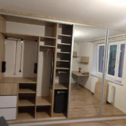 Wnętrze pokoju z dużą szafą wnękową z przesuwnymi drzwiami lustrzanymi, widoczne półki, szuflady i drążki na ubrania, w tle biurko i okno.