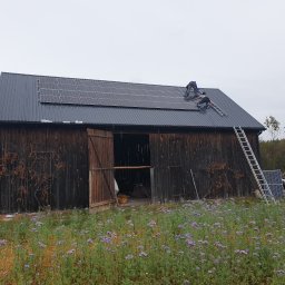 WITVOLT SPÓŁKA Z OGRANICZONĄ ODPOWIEDZIALNOŚCIĄ - Montaż paneli słonecznych na dachu starej, drewnianej stodoły. Dwóch monterów pracuje na dachu, obok drabina. W tle łąka z fioletowymi kwiatami i zabudowania.