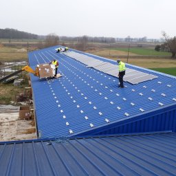 WITVOLT SPÓŁKA Z OGRANICZONĄ ODPOWIEDZIALNOŚCIĄ - Montaż paneli fotowoltaicznych na niebieskim dachu hali magazynowej. Widoczny dźwig podający panele i pracownicy w odblaskowych kamizelkach. Tło: pola i las.