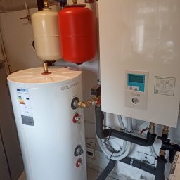 WITVOLT SPÓŁKA Z OGRANICZONĄ ODPOWIEDZIALNOŚCIĄ - Biały zasobnik ciepłej wody Solimpeks 100l z czerwonym i beżowym naczyniem przeponowym oraz pompa ciepła Panasonic Aquarea Smart Cloud z widocznymi rurami.