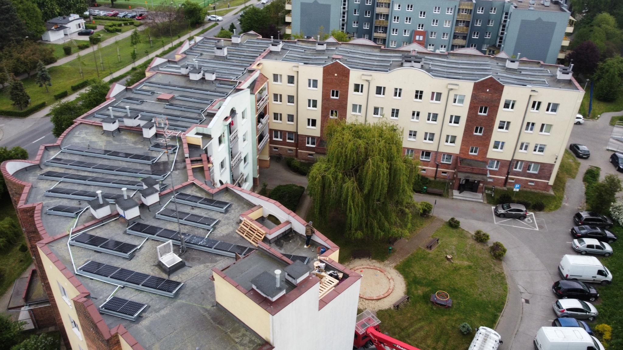 Widok z góry na dach budynku z panelami słonecznymi w trakcie montażu. Część paneli już zamontowana, obok palety i pracownik. W tle bloki mieszkalne i zieleń.