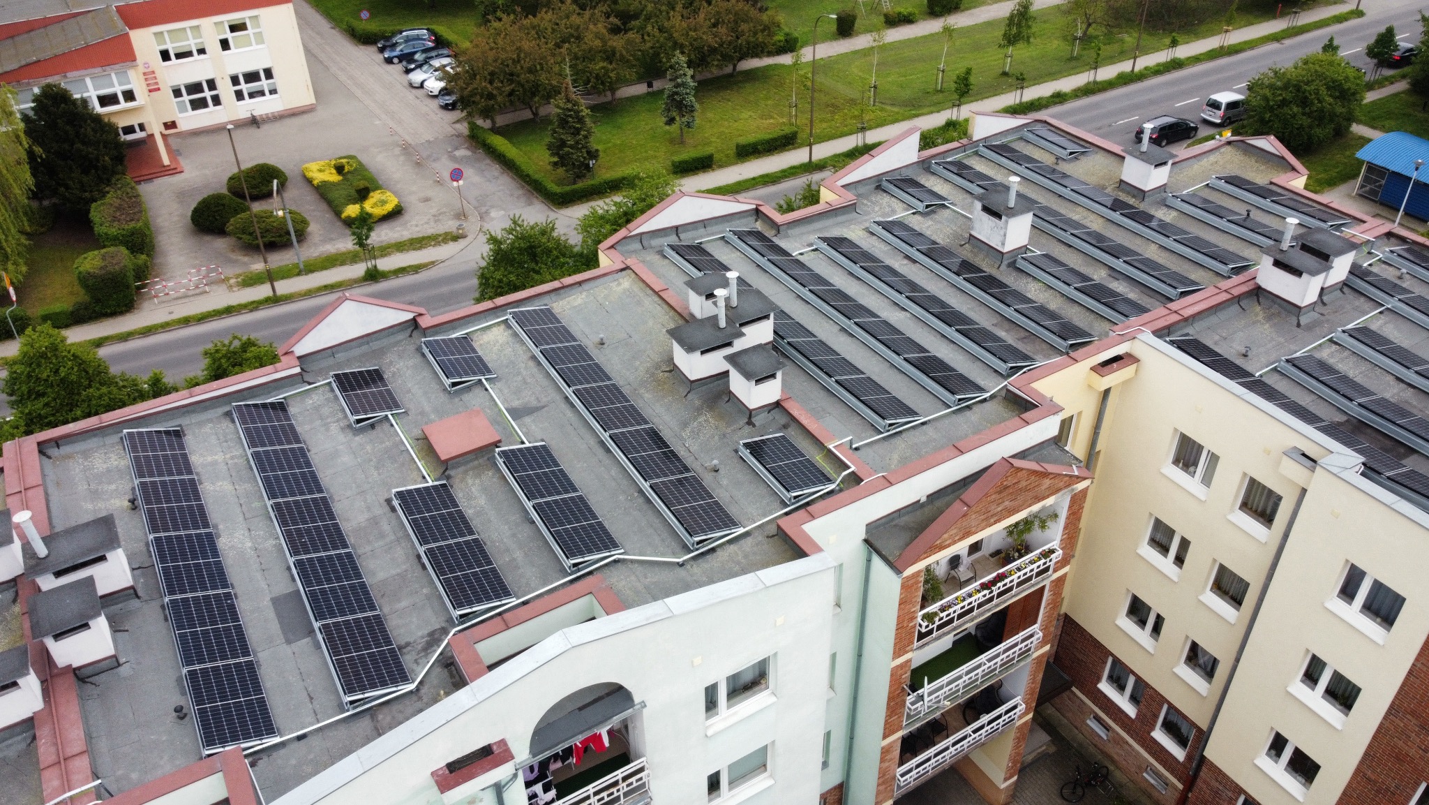Widok z góry na dach budynku z zainstalowanymi panelami fotowoltaicznymi, otoczony zielenią i zabudową miejską. Panele ułożone w rzędach, widoczne kominy wentylacyjne.