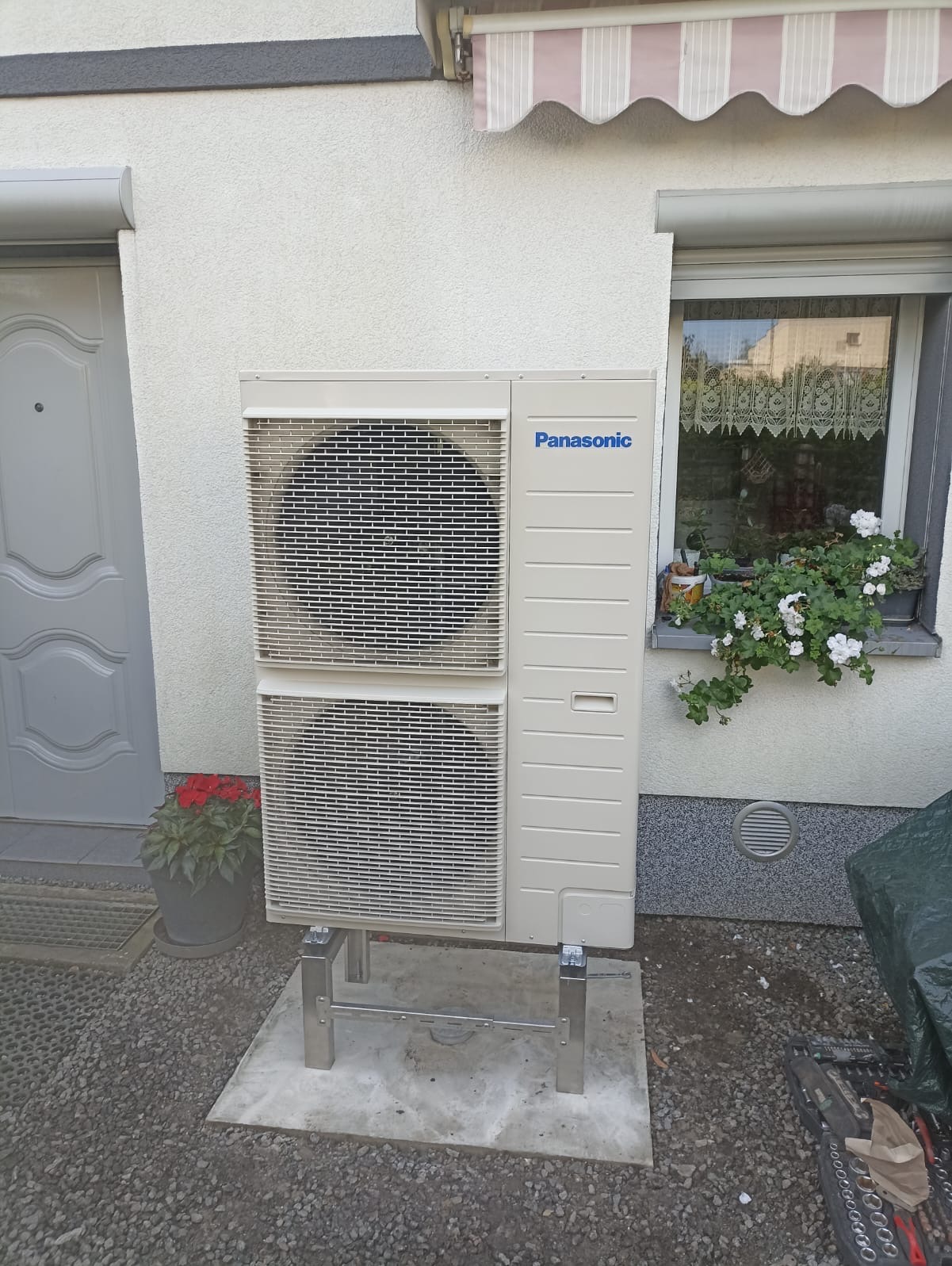 Zewnętrzna jednostka pompy ciepła Panasonic na metalowym stelażu, ustawiona obok drzwi wejściowych i okna z kwiatami, na tle elewacji budynku.
