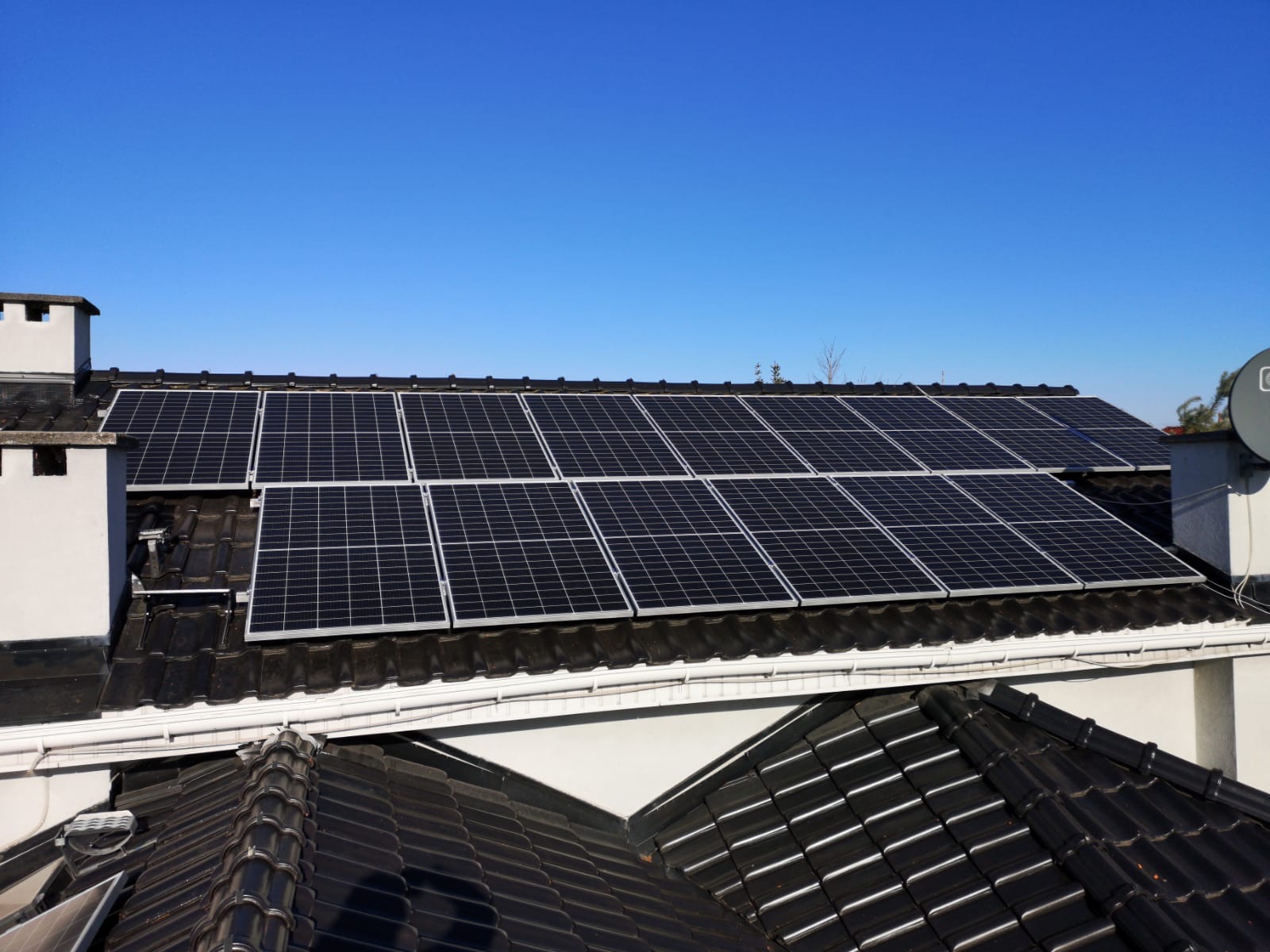 Panele fotowoltaiczne na dachu domu z czarną dachówką, widoczne komin i antena satelitarna. Błękitne niebo w tle. Układ paneli w dwóch rzędach.