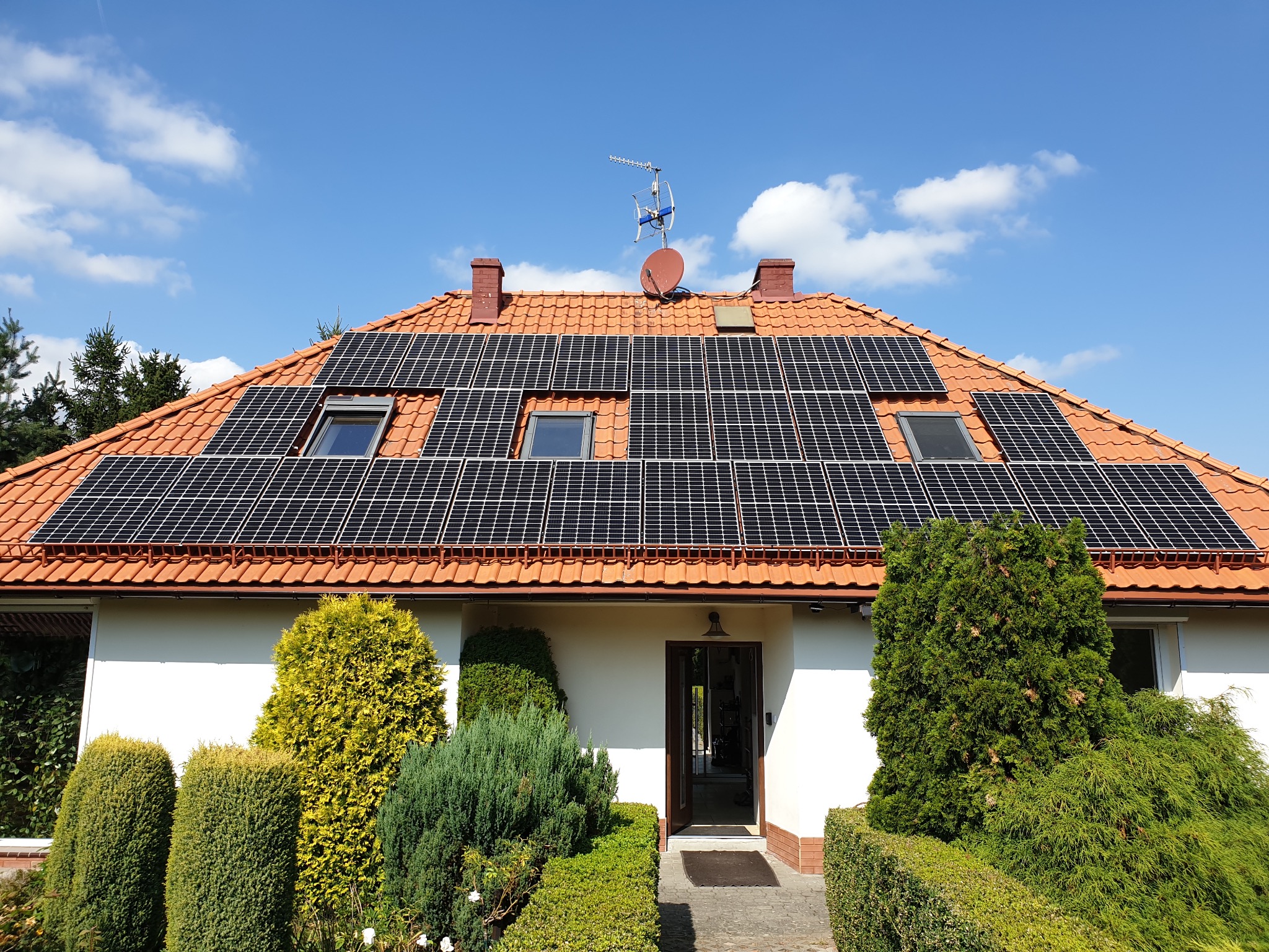 Dom jednorodzinny z panelami słonecznymi na dachu z czerwonej dachówki, widok z poziomu ogrodu, błękitne niebo w tle.