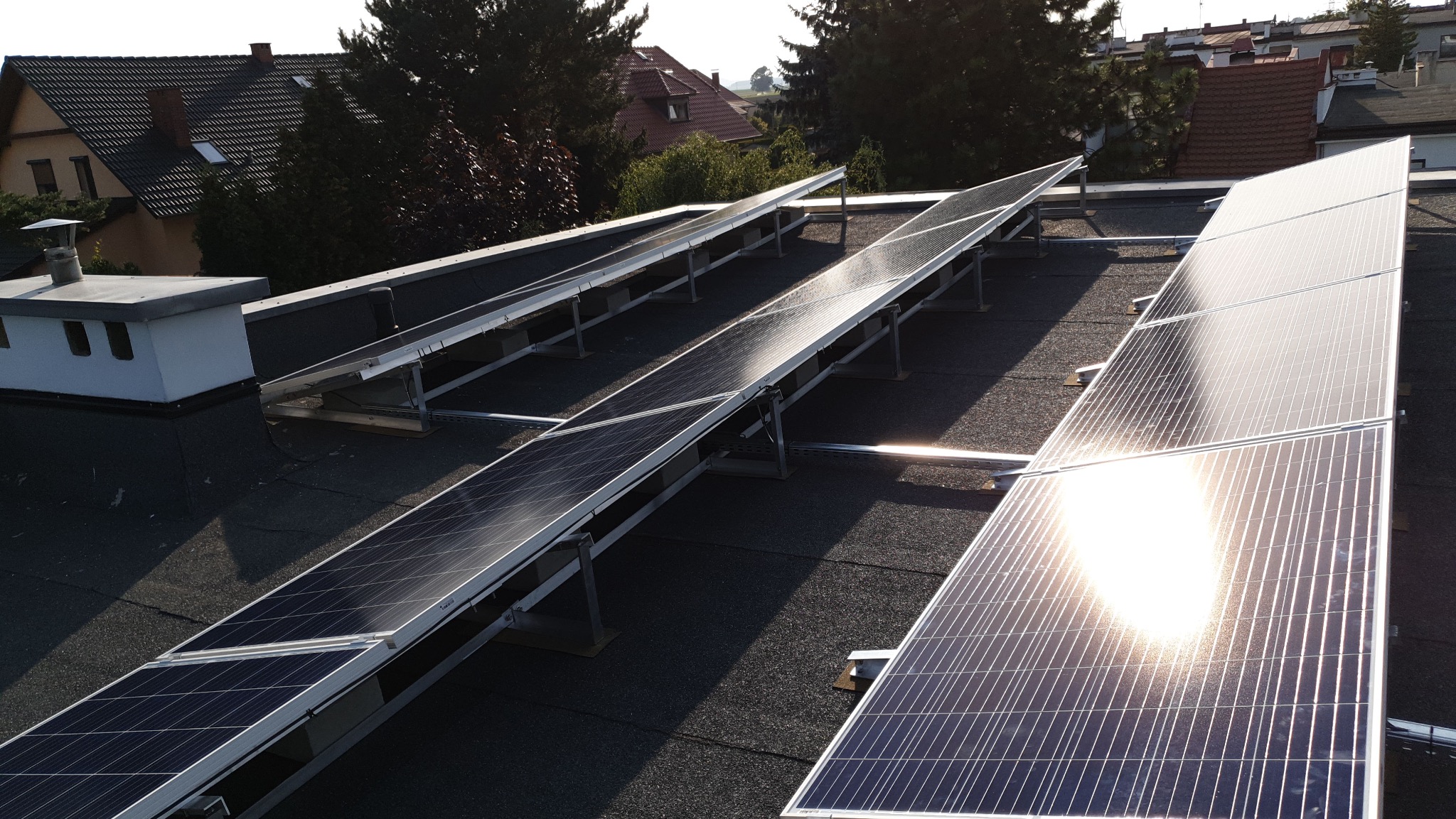 Rząd paneli słonecznych na płaskim dachu z widocznym odbiciem słońca. W tle budynki mieszkalne i drzewa. Instalacja fotowoltaiczna w słoneczny dzień.