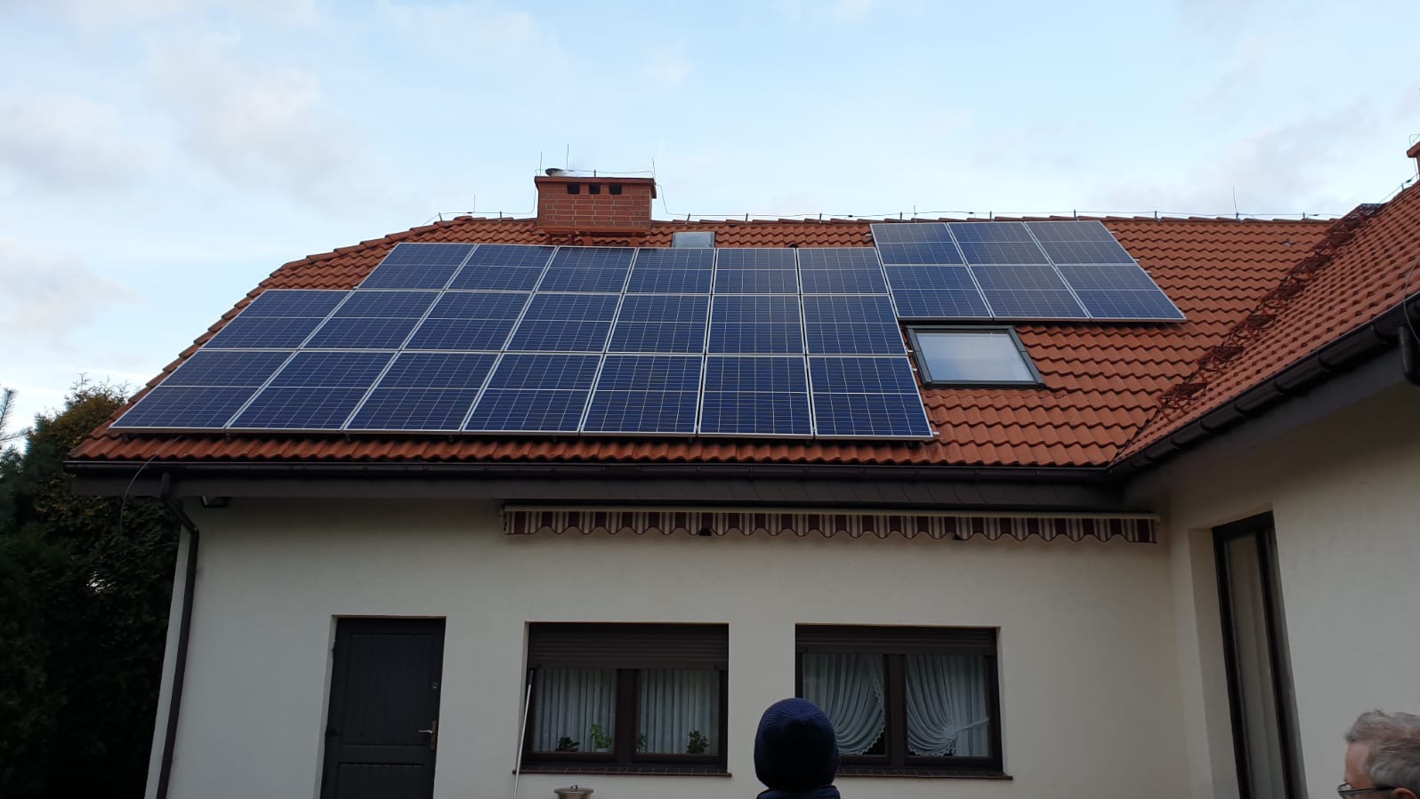 Panele słoneczne na dachu domu z czerwoną dachówką, widoczne okno dachowe i komin. Część budynku w jasnym kolorze z ciemnymi oknami i drzwiami.