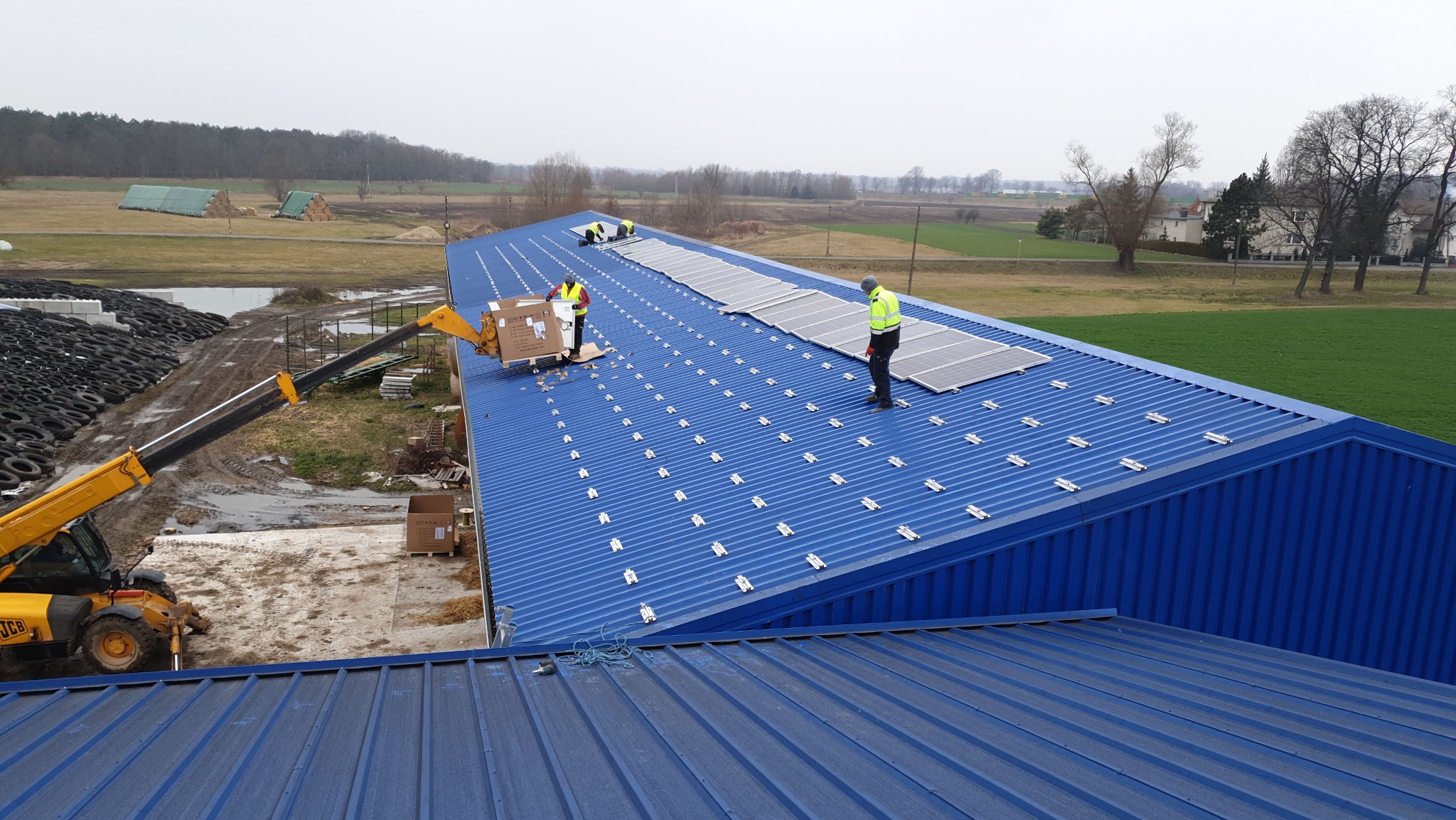 Montaż paneli fotowoltaicznych na niebieskim dachu hali magazynowej. Widoczny dźwig podający panele i pracownicy w odblaskowych kamizelkach. Tło: pola i las.