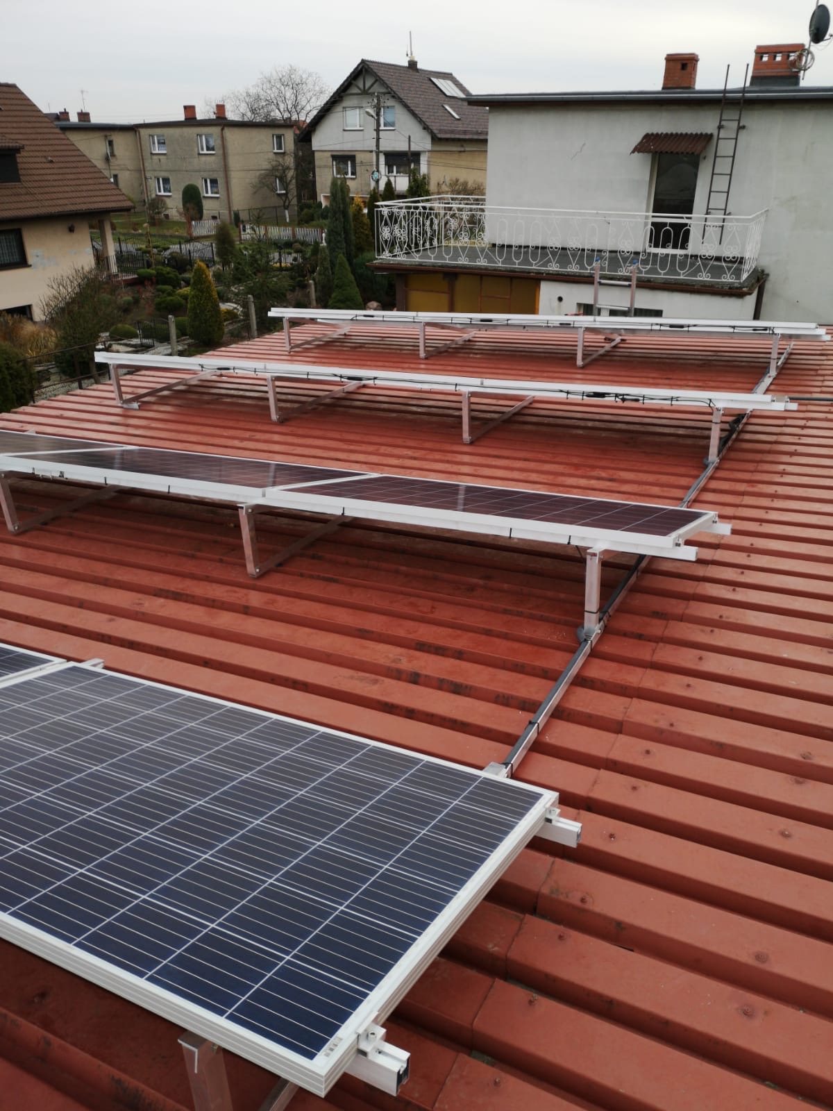 Panele fotowoltaiczne na czerwonym dachu z blachy trapezowej, zamontowane na aluminiowej konstrukcji wsporczej. Widok z góry, w tle zabudowa jednorodzinna.