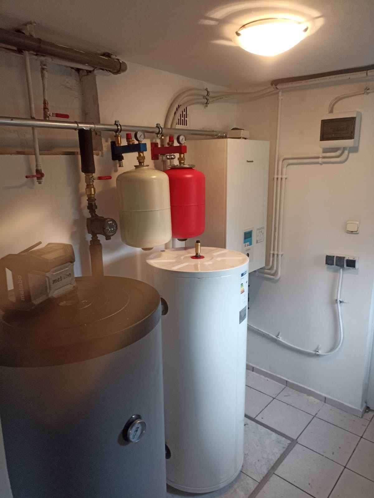 Bojlery w kotłowni: szary i biały, z instalacją hydrauliczną, manometrami i przewodami. Na ścianie biały piec grzewczy i instalacja elektryczna.