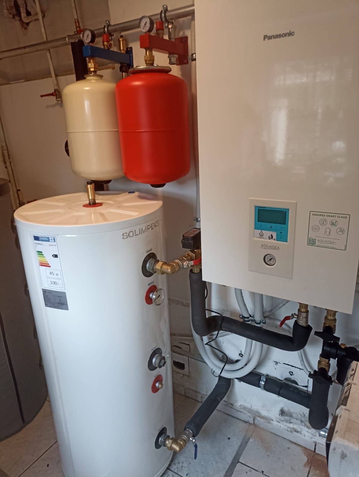 Biały zasobnik ciepłej wody Solimpeks 100l z czerwonym i beżowym naczyniem przeponowym oraz pompa ciepła Panasonic Aquarea Smart Cloud z widocznymi rurami.