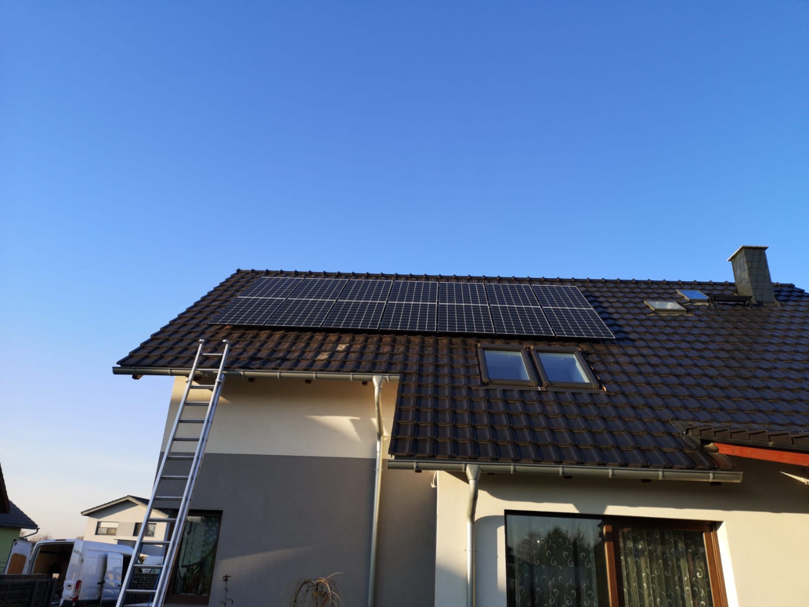 Panele słoneczne na dachu domu z brązową dachówką, drabina oparta o ścianę, błękitne niebo w tle. Widok z dołu.