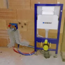 Stelaż podtynkowy Geberit zamontowany przy ścianie z pustaków, widoczne podłączenia wodne i kanalizacyjne, w tle ściana z pustaków.