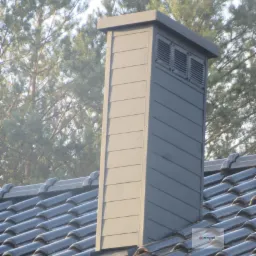 Szary komin wentylacyjny z lamelami na dachu pokrytym ciemną dachówką ceramiczną, na tle drzew iglastych.