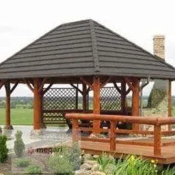 Drewniana altana z ciemnym, skośnym dachem z dachówki, ażurowymi ściankami i kominkiem z cegły w tle zielonego krajobrazu.