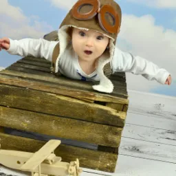 Uśmiechnięty niemowlak w czapce pilota i goglach leży na drewnianej skrzyni, obok drewniany model samolotu na tle błękitnego nieba.