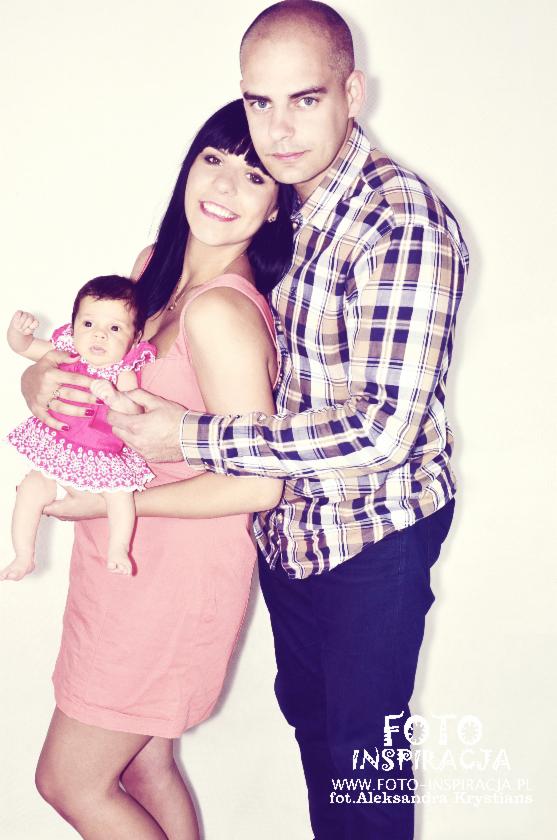 Uśmiechnięta młoda para z niemowlęciem w różowej sukience na białym tle. Kobieta w różowej sukience trzyma dziecko, mężczyzna w koszuli w kratę obejmuje ich ramionami.