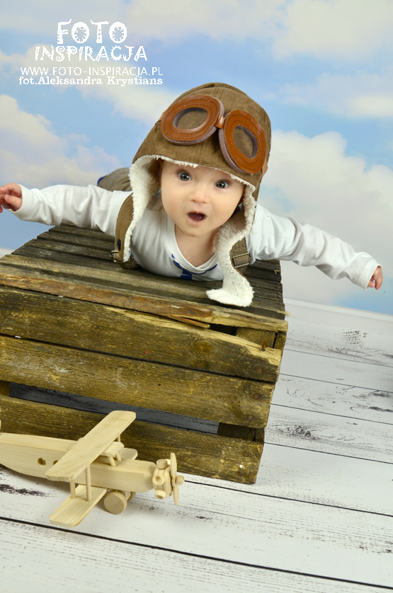 Uśmiechnięty niemowlak w czapce pilota i goglach leży na drewnianej skrzyni, obok drewniany model samolotu na tle błękitnego nieba.