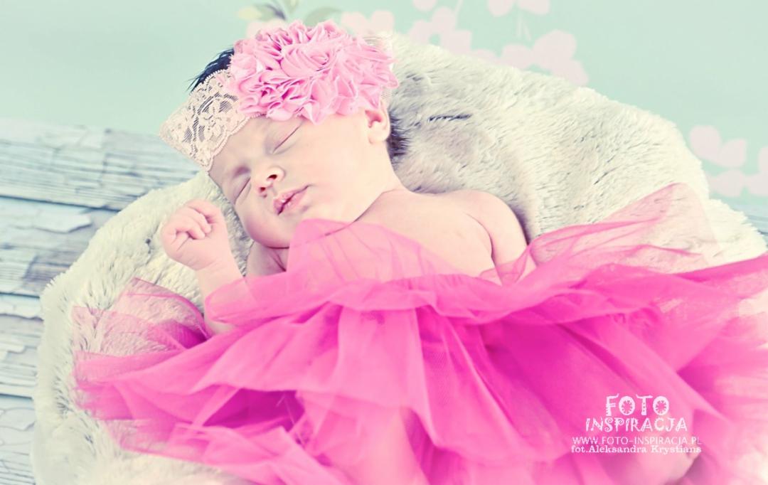 Śpiąca noworodka w różowej tiulowej spódniczce i opasce z kwiatem na tle miękkiego kocyka i pastelowej ściany z kwiatowym wzorem.