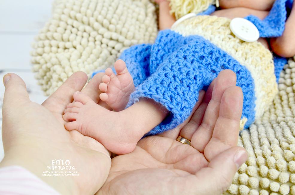 Drobne stópki noworodka w dłoniach rodzica, ubrane w niebieski, dziergany strój, na tle kremowego kocyka.