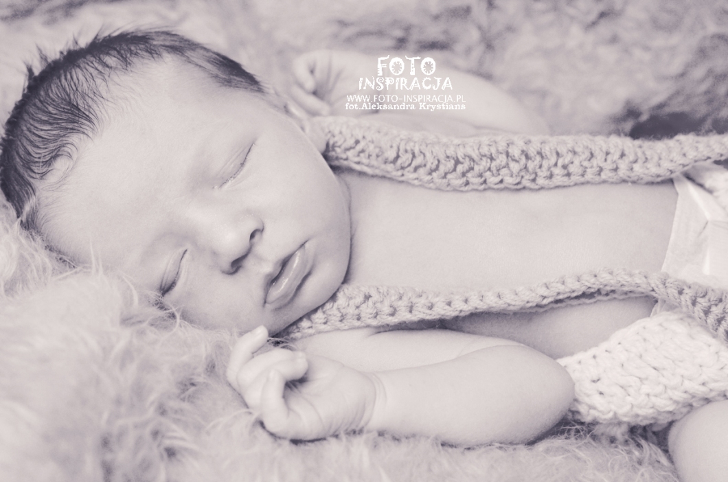 Noworodek śpiący na puszystym tle, ubrany w dziergany strój z szelkami, w czarno-białej tonacji.