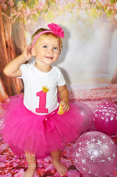 Uśmiechnięta roczna dziewczynka w różowej tiulowej spódniczce i opasce, trzymająca ciastko, pozuje na tle dekoracji urodzinowej z balonami.